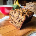 A slice of moist brown butter banana bread filled with chocolate chips on a wooden board