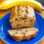 Moist protein banana bread recipe loaf topped with chocolate chips on a blue plate with a fresh banana in the background.