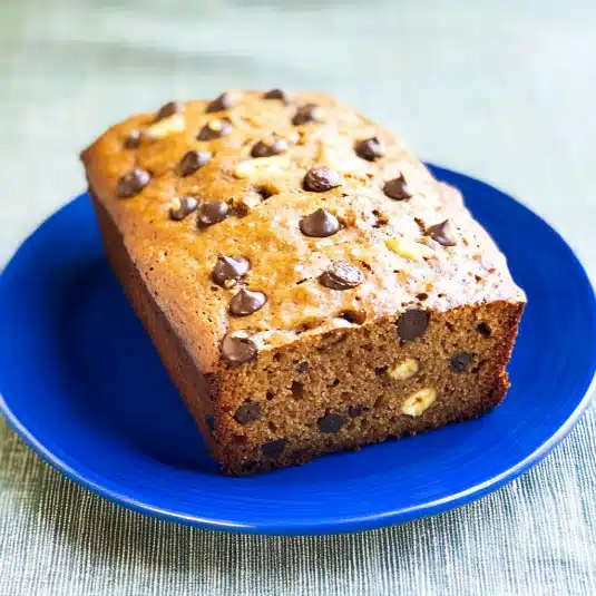 protein banana bread recipe  loaf topped with chocolate chips on a blue plate with a fresh banana in the background.