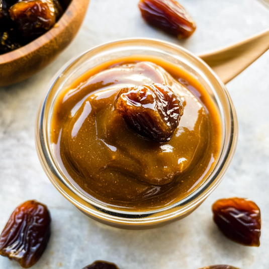 Homemade date caramel in a glass jar topped with a whole date, surrounded by fresh dates on a light background