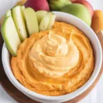 A creamy pumpkin dip in a white bowl topped with whipped spiced pumpkin mixture, surrounded by fresh green apple slices and fall fruits