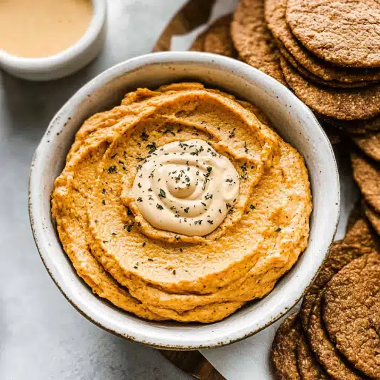 A creamy pumpkin dip in a white bowl topped with whipped spiced pumpkin mixture, surrounded by fresh green apple slices and fall fruits perfect for autumn parties or Thanksgiving desserts.
