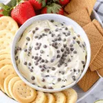 Creamy chocolate chip dip served in a white bowl topped with mini chocolate chips, surrounded by graham crackers, cookies, and fresh strawberries