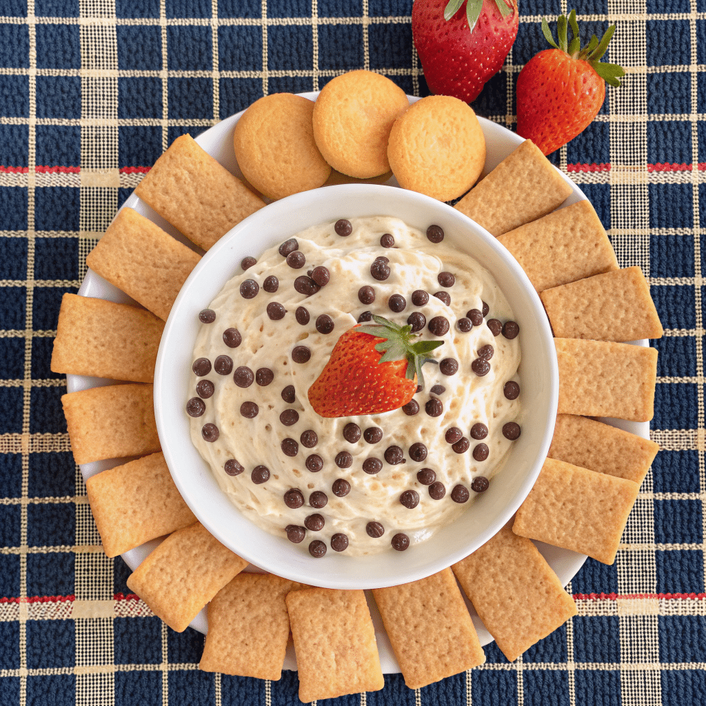 Creamy chocolate chip dip served in a white bowl topped with mini chocolate chips, surrounded by graham crackers, cookies, and fresh strawberries